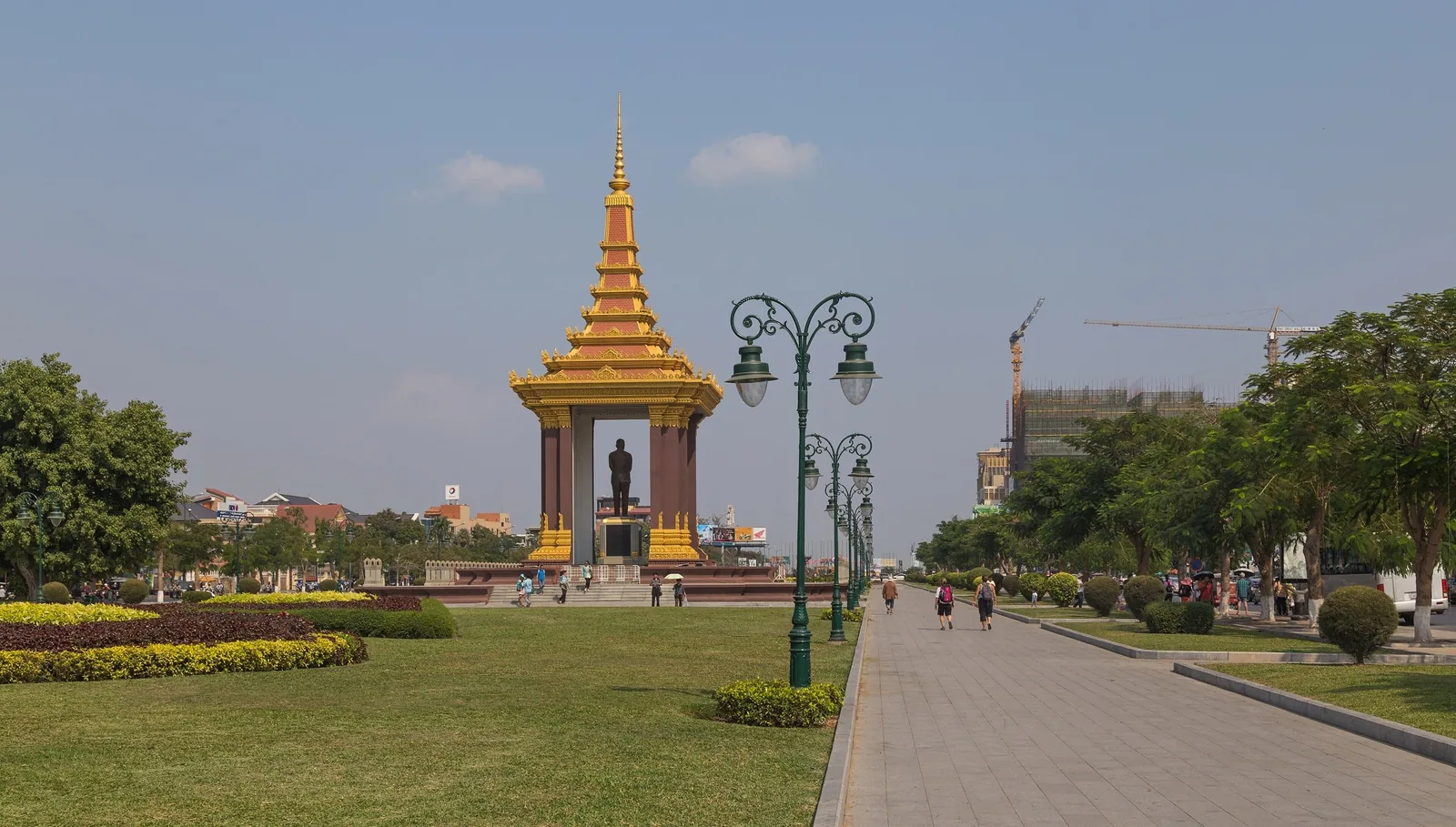 A gilded pavilion housing a statue of a former king stands along a landscaped park path in Phnom Penh.