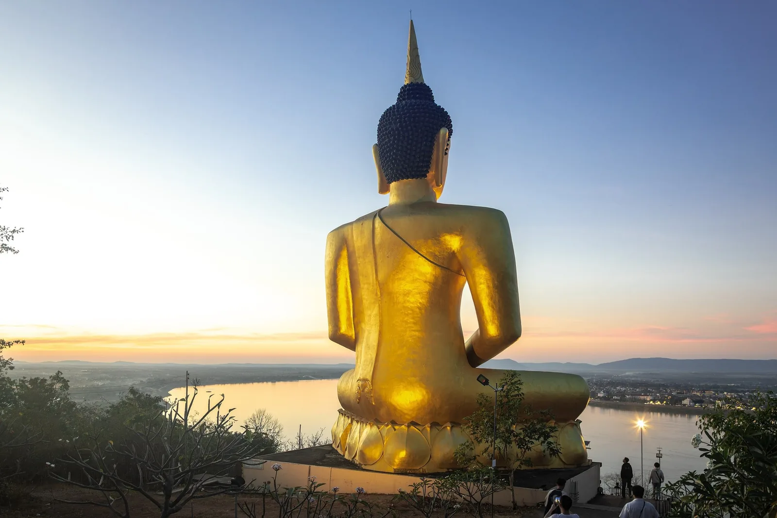 Golden Buddha statue seen from behind atop a hill overlooking the Mekong River in Pakse at sunset.