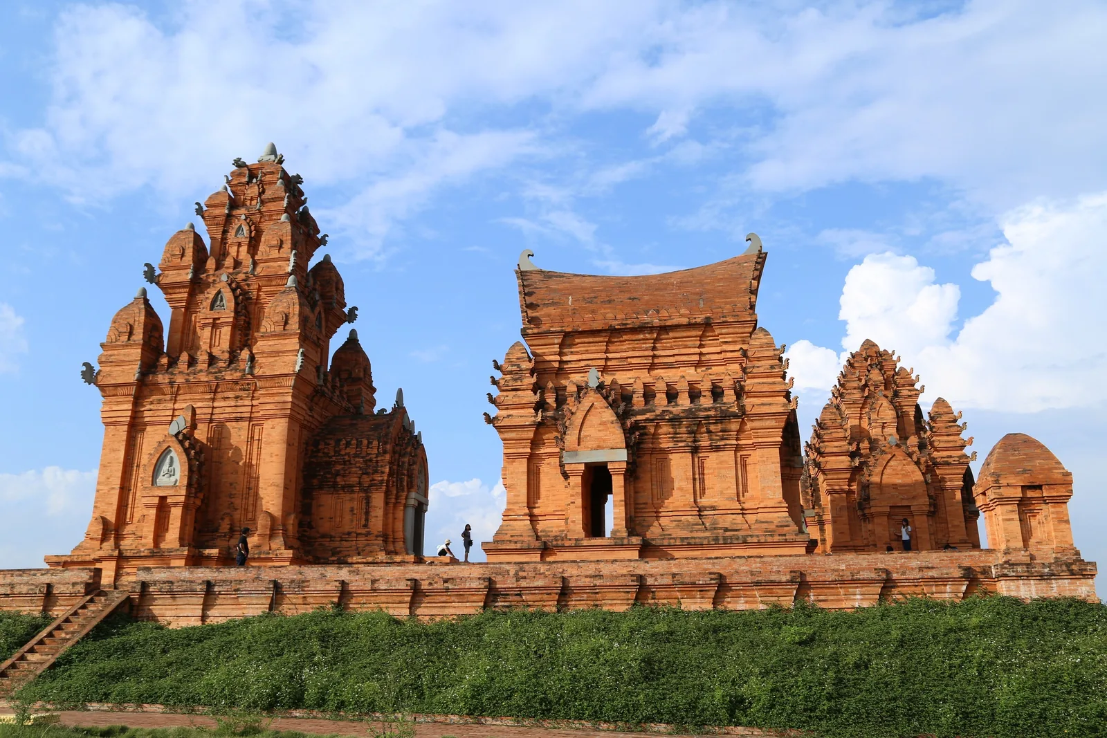 Ancient red brick Cham towers stand atop a green hillside under a bright blue sky in Vietnam.