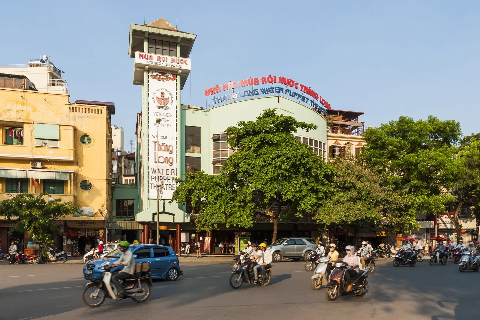 Motorbikes and cars pass the Thang Long Water Puppet Theatre building in Hanoi under a clear sky.