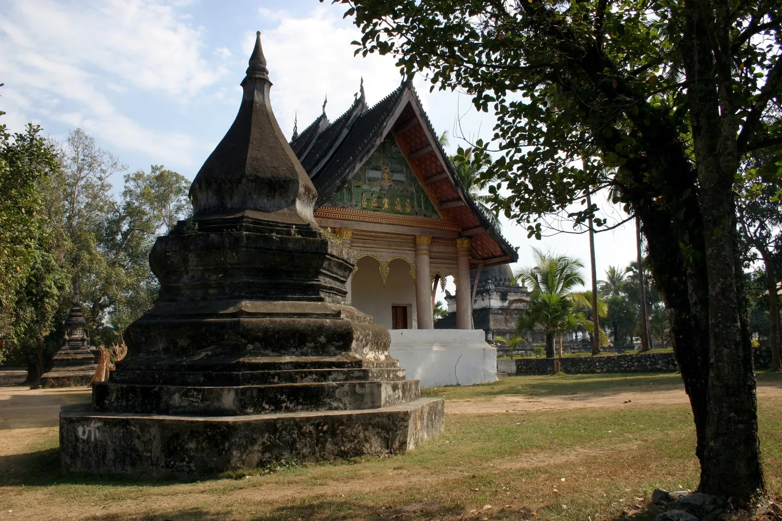 A weathered stone stupa stands before a temple hall with an ornate gabled roof at Wat Aham in Luang Prabang.