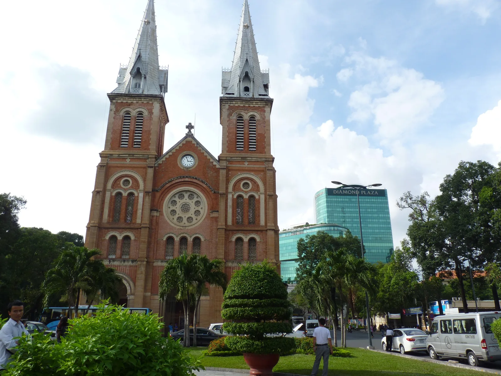 The red-brick twin spires of Notre Dame Cathedral rise above palm trees with a modern glass tower behind in Ho Chi Minh City.