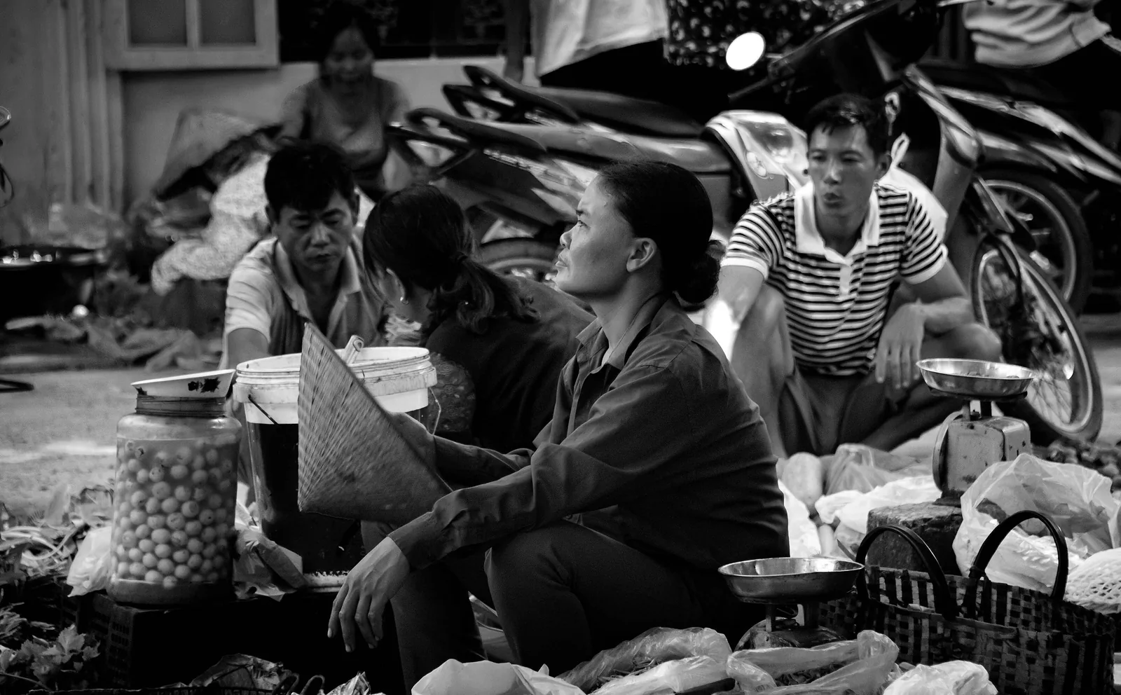 Vendors and shoppers sit among baskets and jars at a street market in Hanoi, Vietnam.