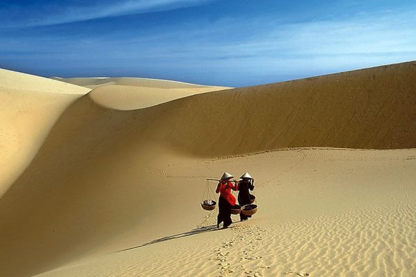 Two people carrying baskets across the ridge of a pale sand dune near Mui Ne
